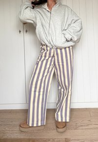 Siva jopa jopa z zadrgo z spuščenimi rameni, kombinirana z širokimi hlačami z navpičnimi kremnimi in vijoličnimi črtami. Model nosi bež obutev.