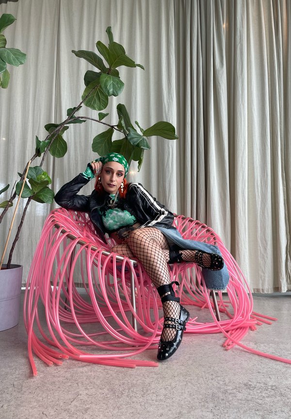 Rosa plaistigh aidhm le dearaidh tonnta, samhail i seaicéad lisa dubh agus stocaí iasc, t-uige duille ghlas, ina sui i measc duilleoga móra ghlas.