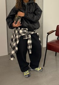 Personne portant une veste en cuir noire, un pantalon noir, une chemise à carreaux nouée autour de la taille, des baskets jaunes et grises, tenant un sac à main à motifs à l'intérieur.