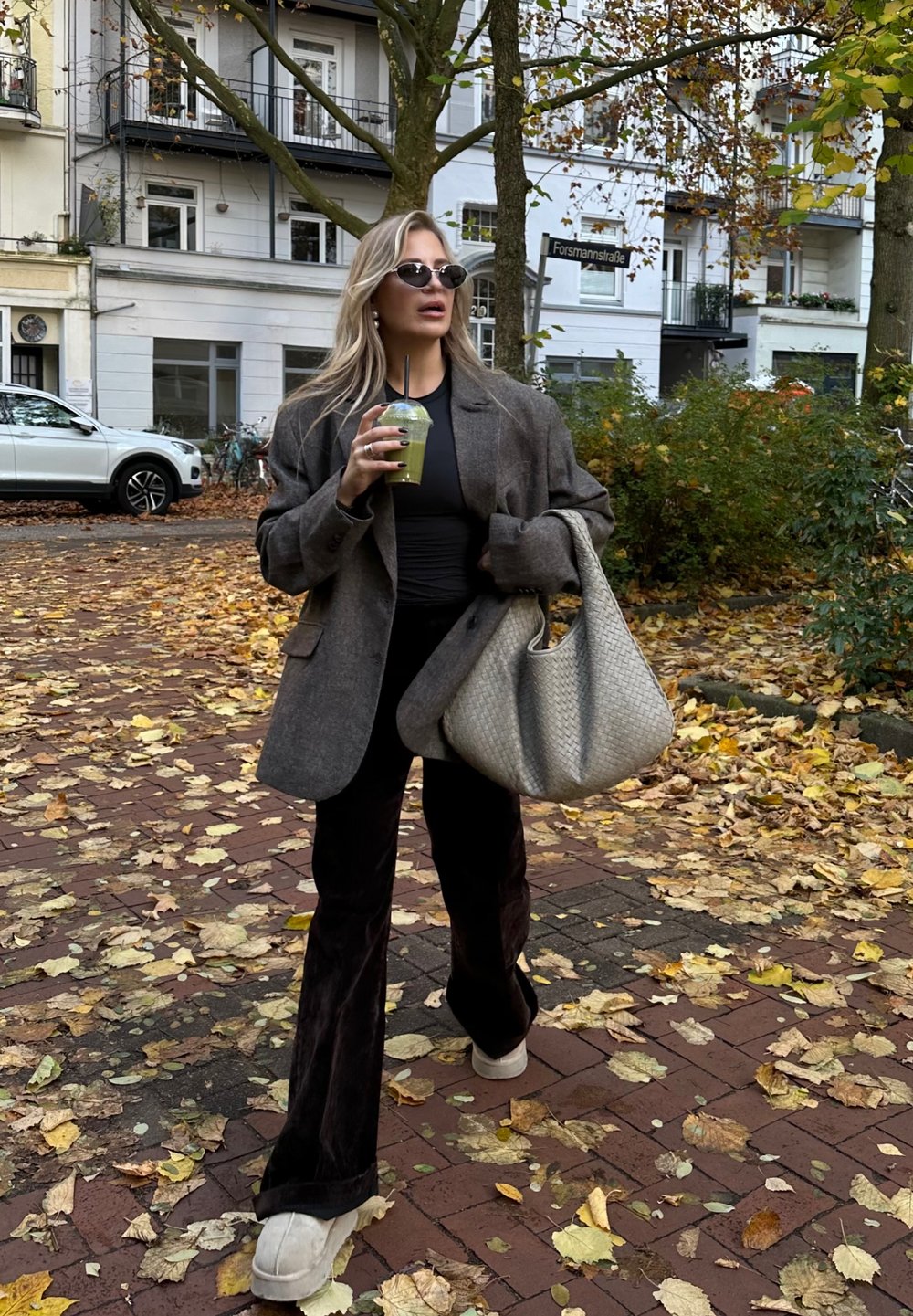 Blazer cinzento oversized, blusa preta e calças escuras de boca de sino. Segurando uma bebida verde. Grande mala tecida bege. Folhas de outono no chão.