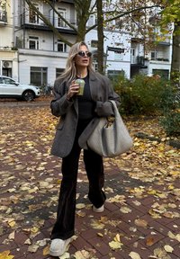 Blazer cinzento oversized, blusa preta e calças escuras de boca de sino. Segurando uma bebida verde. Grande mala tecida bege. Folhas de outono no chão.