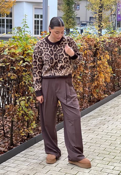 Suéter de estampado de leopardo en tonos marrón y beige, combinado con pantalones holgados marrones y zapatos slip-on beige, con un fondo de hojas.