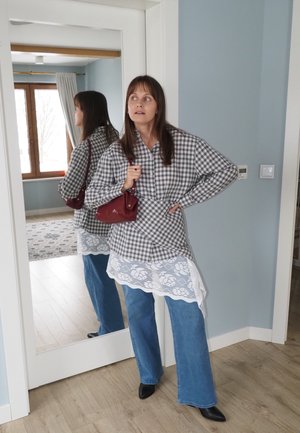 Mujer con camisa de cuadros, vestido con encaje, vaqueros azules y zapatos negros, sosteniendo un bolso rojo, de pie junto a un espejo de cuerpo entero en una habitación azul clara.