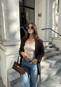 Palton din lână maro, bluză albă cu dungi, blugi ușori cu talie înaltă de culoare albastră, geantă din suede maro cu detalii aurii și ochelari de soare închiși.