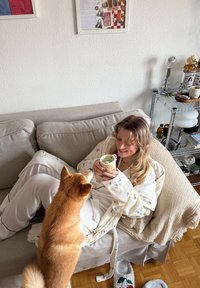 Une femme en robe légère à motifs tient une tasse à motifs. Un Shiba Inu est tout près. Le canapé est beige, avec un sol en bois visible.