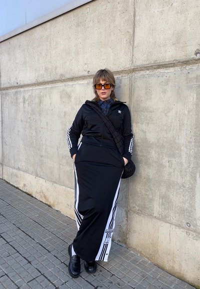 Chaqueta de chándal negra y falda maxi con rayas blancas, combinadas con zapatos negros. La chaqueta tiene cremallera y un logo; las gafas de sol completan el look.
