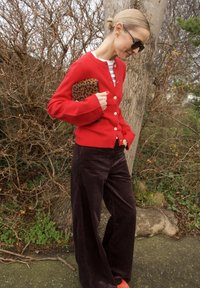 Cardigan rouge avec des boutons dorés, associé à un pantalon large en velours côtelé marron foncé. Tient une pochette imprimée léopard, sur un fond naturel.