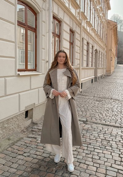 Mujer de cabello largo con abrigo trench beige, suéter de punto blanco, pantalones anchos y zapatillas, de pie en una calle adoquinada junto a las ventanas altas de un edificio.