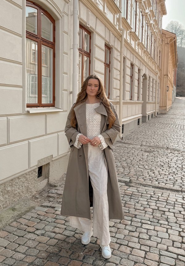 Vrouw met lang haar, gekleed in een beige trenchcoat, witte gebreide trui, wijde broek en sneakers, staand op een keienstraat bij hoge ramen van een gebouw.