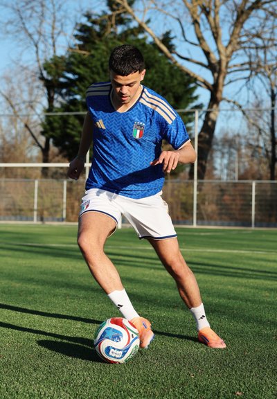 Joven jugador de fútbol masculino con camiseta azul de Italia controlando un balón de fútbol en un campo de césped con árboles y una cerca en el fondo.