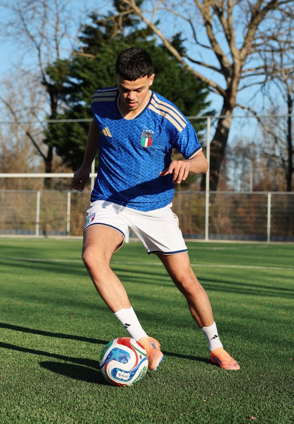 Jeune joueur de football masculin portant un maillot bleu de l'Italie, contrôlant un ballon de football sur un terrain en herbe avec des arbres et une clôture en arrière-plan.