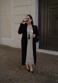 Cappotto lungo nero su un vestito in tessuto cremoso con bordo in pizzo, accessorato con una collana a pendente e una borsa nera, posato su una pavimentazione in ciottoli.