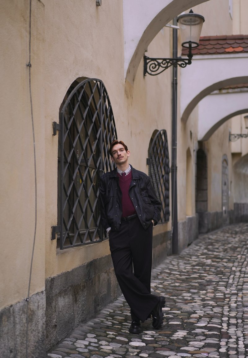 Jeune homme en veste noire et pantalon large appuyé contre un mur beige avec des fenêtres cintrées et une rue pavée sous des arches.