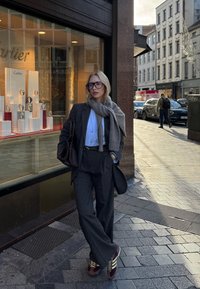 Gråt uldjakkesæt med løs pasform, lyseblå skjorte og gråt tørklæde. Brun håndtaske og bordeaux sneakers med beige accenter. Butiksvindue viser ure.