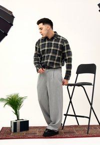 Homme en chemise à carreaux et pantalon gris debout sur un tapis à motifs près d'une chaise haute noire, d'une petite plante et d'une radio vintage.