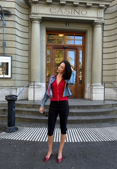 Mujer con top rojo, pantalones negros cortos y chaqueta vaquera de pie, tocándose el pelo frente a un edificio con la etiqueta "CASINO" con escalones de piedra.