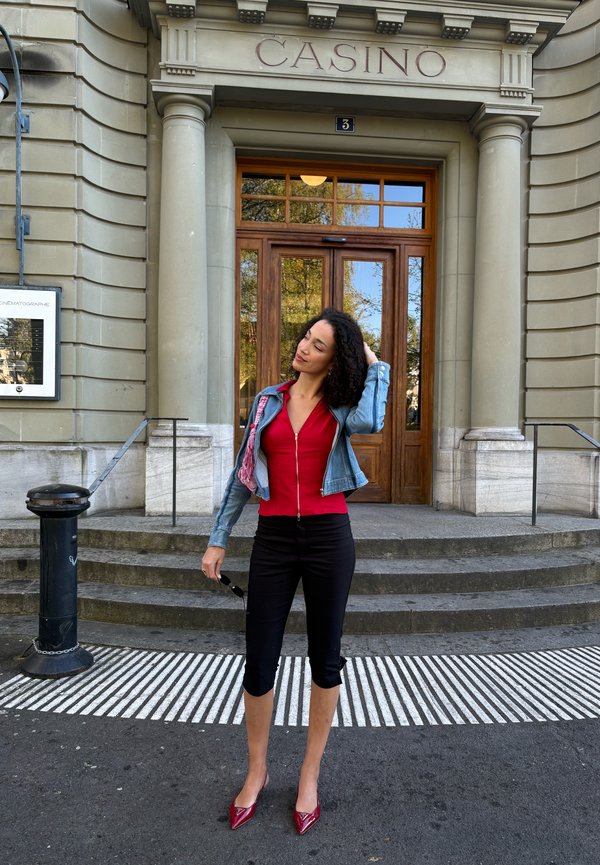 Woman in red top, black cropped pants, and denim jacket standing, touching hair in front of a building labeled "CASINO" with stone steps.