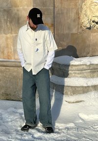Homme portant une chemise à manches longues blanche sous une chemise à manches courtes, un jean bleu, une casquette noire et des baskets, debout sur la neige près d'une structure en pierre.