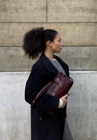 Frau mit lockigem Pferdeschwanz, die einen schwarzen Mantel und ein graues Shirt trägt, ein dunkelbraunes Lederhandtasche haltend, seitlich gegen eine strukturierte Steinwand stehend.