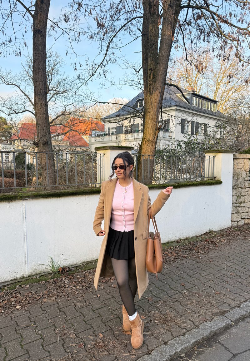 Long manteau beige sur un cardigan rose, jupe noire et collants noirs. Sac marron et bottes camel. Décor extérieur avec des arbres et une maison.
