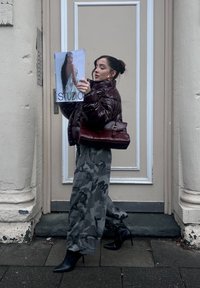 Maroon padded jacket, camouflage trousers, black pointed heels. Holding a magazine featuring "STUDIO" with a model on the cover.