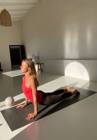 Naine punases topis ja mustades retuusides teeb Cobra jooga poosi mustal mattidel päikesepaistelises minimalistlikus toas, kus on painduv riputustuli ja tool.