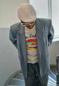 Blazer azzurro chiaro su una maglietta bianca con un motivo di pesci colorati, indossato con jeans di denim. Un cappello chiaro e occhiali da sole completano il look.