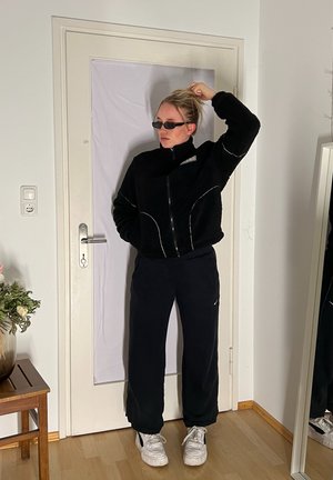 Chaqueta de forro polar negra con costuras blancas en contraste, cierre de cremallera, cuello alto; pantalones negros con bolsillos laterales; zapatillas blancas.