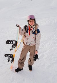 Jeune femme en tenue de snowboard tenant un snowboard, debout sur une pente enneigée avec des montagnes couvertes de neige en arrière-plan.
