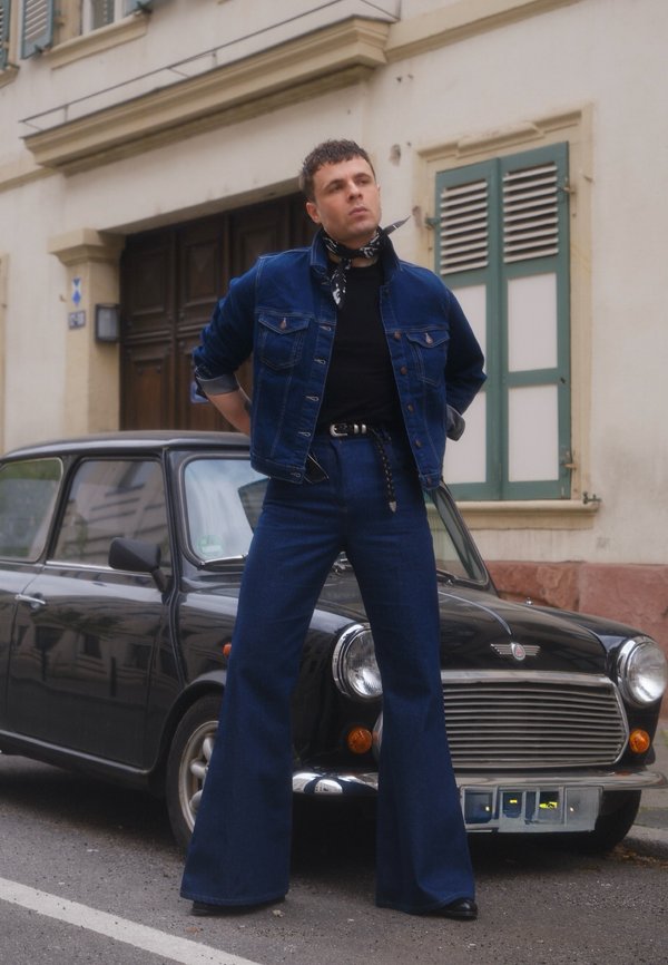 Uomo con giacca di jeans e jeans a zampa si trova con le mani dietro la schiena davanti a una Mini Cooper vintage nera in una via della città.