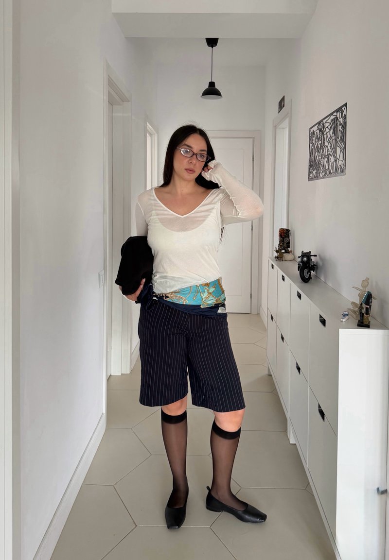 Femme avec des lunettes, top blanc, écharpe bleue à motifs, short à fines rayures et chaussettes transparentes montantes jusqu'aux genoux, debout dans un couloir aux murs et placards blancs.