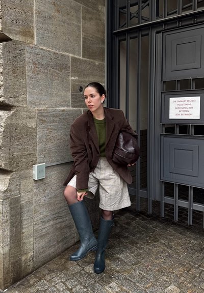 Mujer con blazer marrón, suéter verde, pantalones cortos de cuadros y botas de lluvia azules ajustándose la bota cerca de una puerta metálica gris y una pared de piedra.
