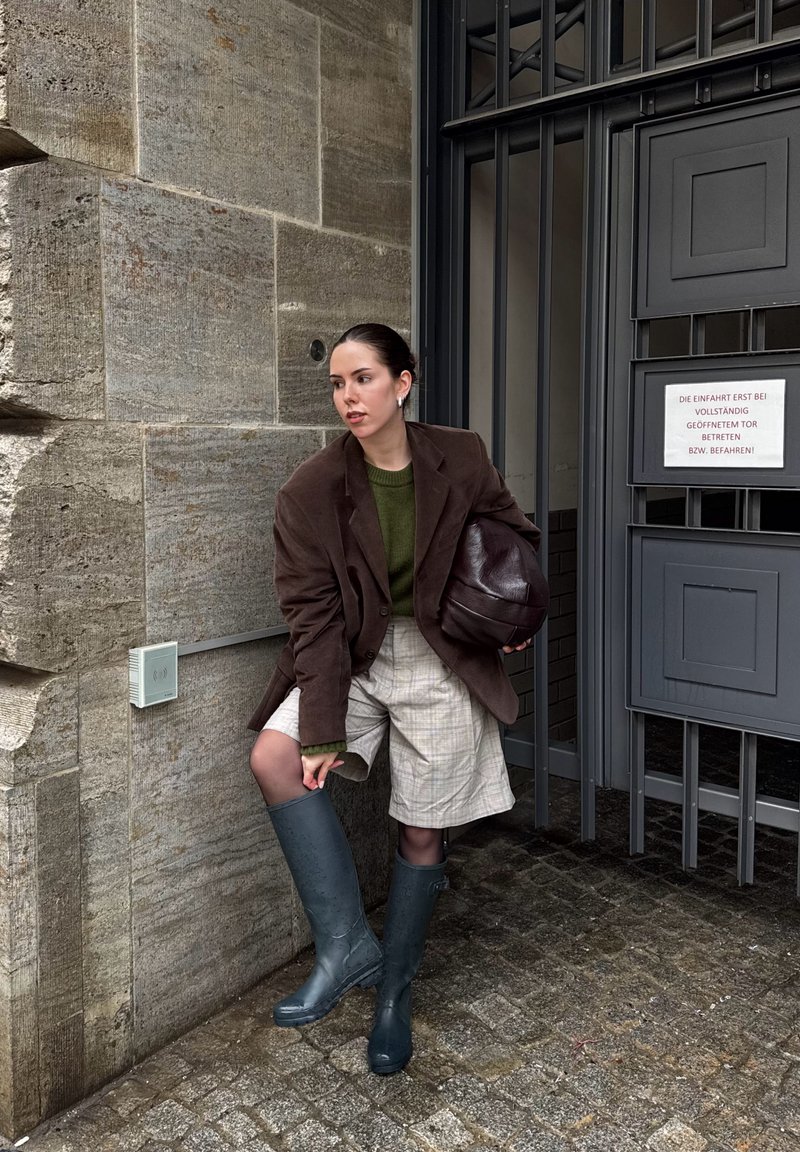 Žena v hnedom saku, zelenom svetri, károvaných krátkych nohaviciach a modrých gumákoch si upravuje topánku pri šedej kovovej bráne a kamennej stene.