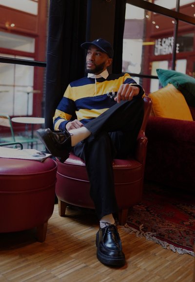 Hombre sentado en un sillón burdeos, vistiendo una camiseta de rugby a rayas amarillas y marinas, pantalones negros y zapatos negros. Suelo de madera, ambiente interior.