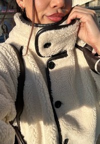 Woman wearing a cream fleece jacket with black buttons and trim, touching the collar, with glossy lips and small gold hoop earrings visible.