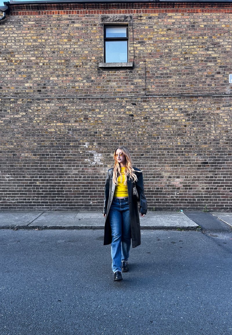 Femme aux longs cheveux blonds portant un manteau noir, un haut jaune et un jean marche sur le trottoir devant un mur de briques marron avec une petite fenêtre.