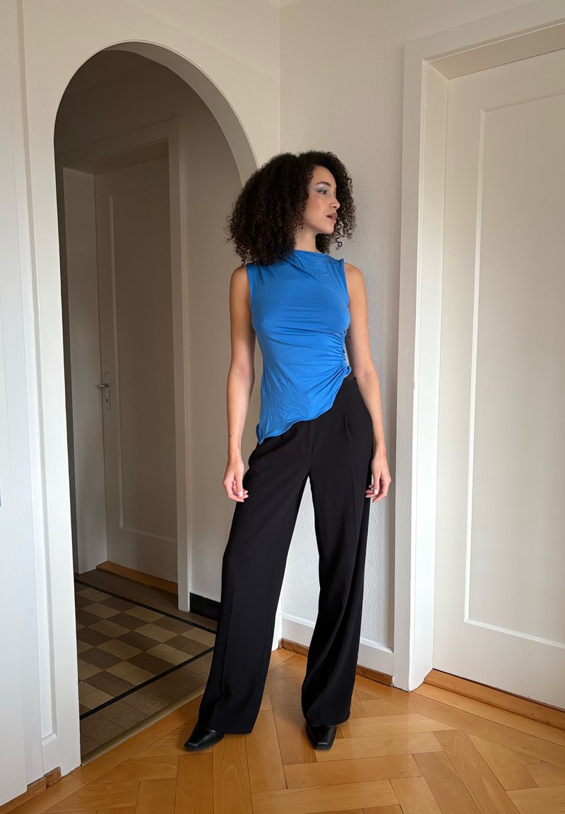 Haut bleu sans manches avec détail torsadé, associé à un pantalon noir à jambes larges. Tissu lisse, silhouette ajustée et chaussures pointues noires.