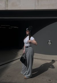 Camiseta blanca ajustada, pantalones de chándal grises de pierna ancha con una cinturilla "Supreme", gorro negro, gafas de sol y un bolso negro, de pie sobre el concreto.