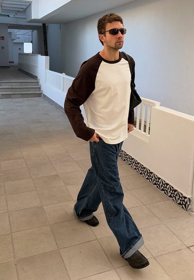 Hombre con gafas de sol, camisa de manga larga en blanco y negro, vaqueros anchos azules y zapatos negros caminando en el interior por un pasillo con baldosas.