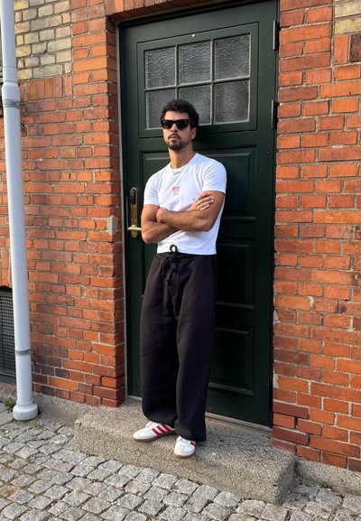 Hombre con gafas de sol, camiseta blanca, pantalones negros y zapatillas blancas de pie con los brazos cruzados frente a una puerta verde oscura en un edificio de ladrillo.