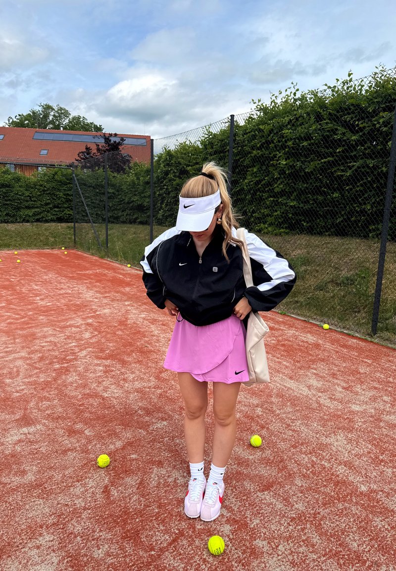 Jeune femme portant une tenue de tennis se tient sur un court en terre battue entourée de balles de tennis, avec une haie verte et une clôture en filet en arrière-plan.