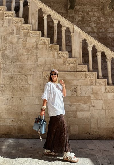Camisa blanca de gran tamaño, falda marrón texturizada, bolso azul, zapatillas con estampado; de pie junto a una escalera de piedra con columnas decorativas.