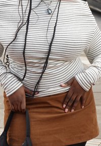 Camisola de manga comprida às riscas em branco e preto, combinada com uma saia castanha de camurça. Apresenta uma textura subtil, com uma fita preta cuidadosamente atada e mãos apoiadas nos quadris.