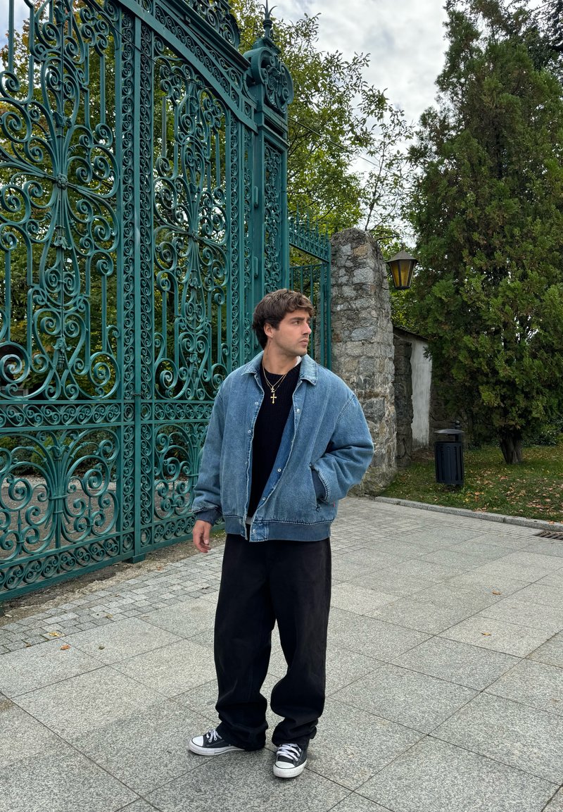 Τζάκετ τζιν, άνετη εφαρμογή, ανοιχτό μπλε με αντίθετο γιακά. Μαύρη πλεκτή μπλούζα, φαρδιά μαύρα παντελόνια και γκρι αθλητικά παπούτσια. Επιτελούμενη πράσινη πύλη στο φόντο.