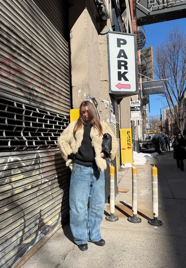 Kvinde iført oversized blå jeans, sort hættetrøje og beige pelsjakke står op ad en bygning med et "PARK"-skilt og en lukket metalklap.