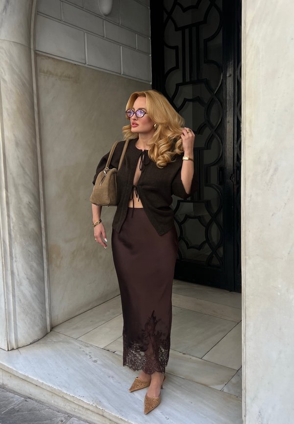 Femme blonde portant des lunettes, un cardigan court foncé, une jupe marron bordée de dentelle et des talons pointus, debout sur des marches en marbre près d'une porte noire.