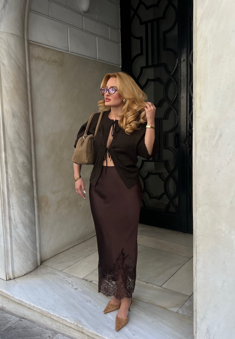 Femme blonde portant des lunettes, un cardigan court foncé, une jupe marron bordée de dentelle et des talons pointus, debout sur des marches en marbre près d'une porte noire.