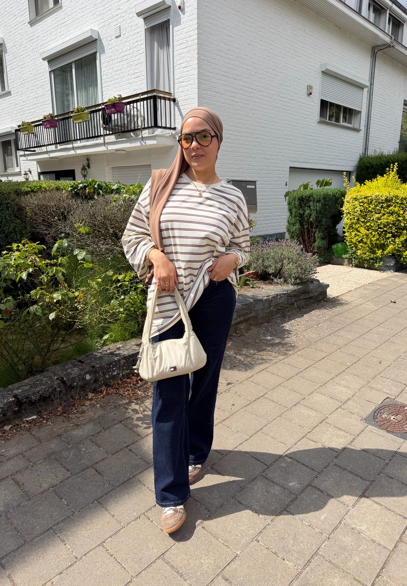 Femme portant un foulard brun sur la tête et des lunettes, une longue chemise à rayures et un pantalon foncé, debout sur un trottoir pavé près de buissons verts et de bâtiments blancs.