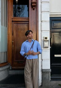 Lichtblauwe button-up shirt, beige gestreepte wijde broek, zilveren sieraden, staande bij een houten deur met nummer 200, intercom op de muur.