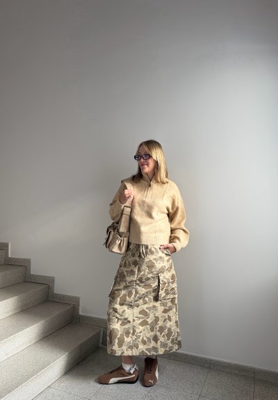 Suéter de punto beige con media cremallera, combinado con una falda de camuflaje hasta la rodilla y zapatillas marrones. Un bolso marrón cuelga del hombro.